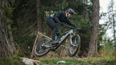 Se ve a un hombre en una bicicleta eléctrica Cube Stereo Hybrid gris saltando por una pendiente en el bosque.