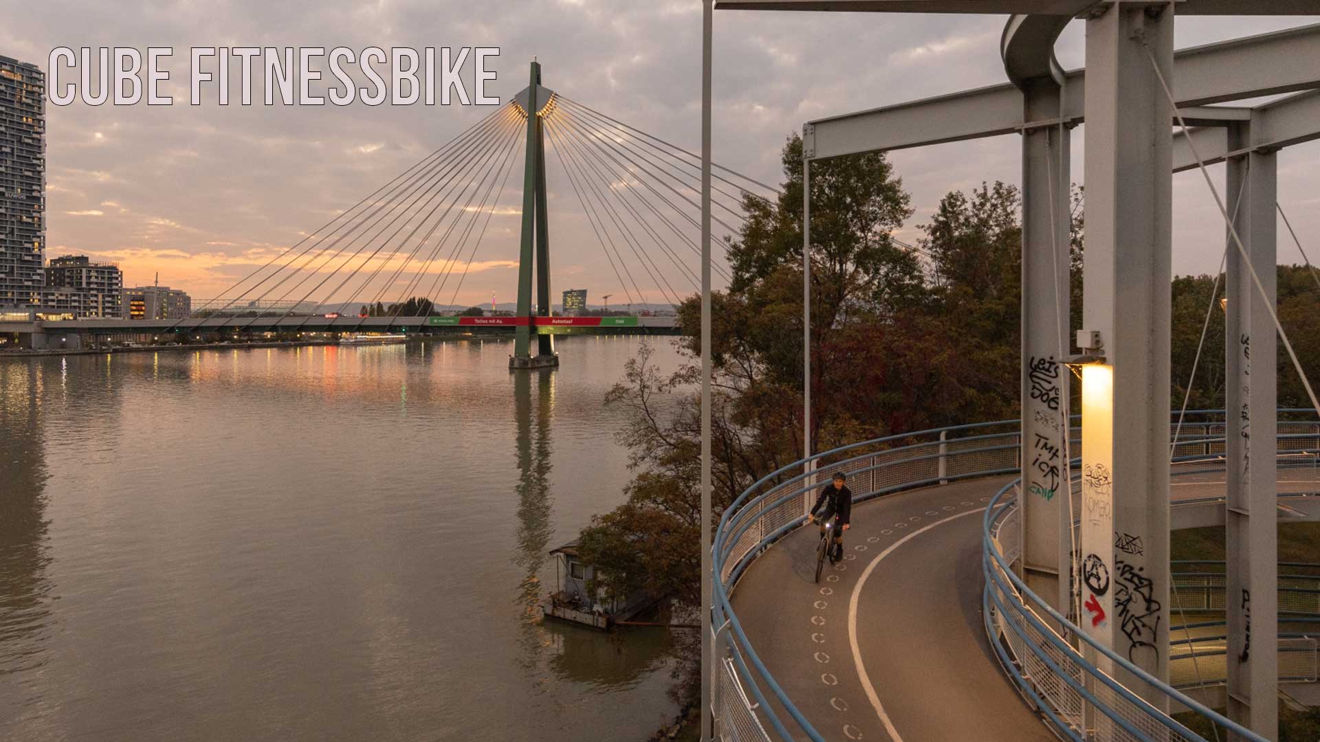 Se ve una ciudad a orillas de un río. En primer plano hay una ciclista que circula por el carril bici al atardecer. La imagen lleva el título «Cube Fitnessbike».