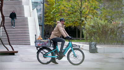 Se ve a un hombre en una bicicleta eléctrica de carga Cube Longtail Hybrid azul, que circula por el centro de la ciudad con alimentos almacenados.