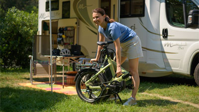 Una mujer está delante de una autocaravana, delante de ella hay una bicicleta plegable verde.