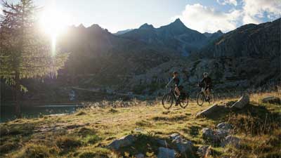 Una pareja recorre un sendero con sus bicicletas de montaña. Al fondo se ven las montañas, detrás de las cuales asoma el sol.