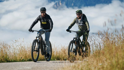 Dos ciclistas circulan en sus bicicletas eléctricas de montaña por un carril bici. Al fondo se ven las montañas.