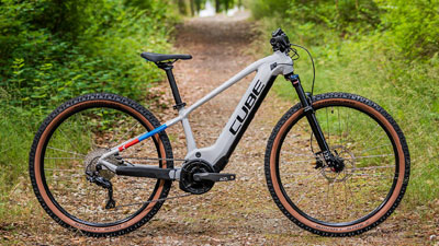 Se ve una bicicleta eléctrica infantil Cube blanca, con pequeños detalles en azul y rojo. Está parada en un camino forestal cubierto de vegetación.
