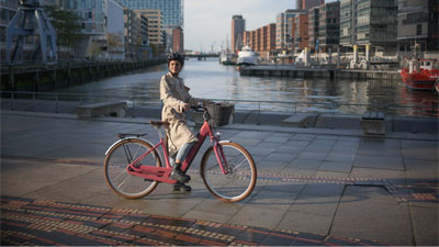 Se ve a una mujer montada en una bicicleta eléctrica Ella Hybrid de color rosa. Está circulando junto a un río. Al fondo se ve una ciudad.