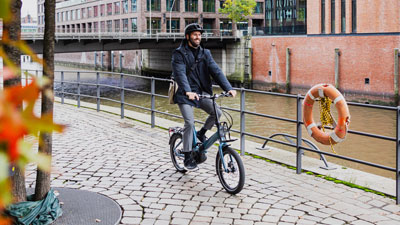 Un hombre va en su bicicleta plegable azul, con casas y un río al fondo.