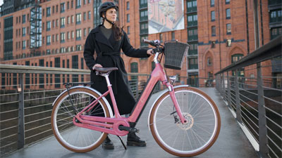 Se ve a una mujer detrás de una bicicleta eléctrica rosa Cube Ella Hybrid. Al fondo hay un edificio.