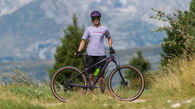 Se ve a una mujer con ropa de ciclista morada, delante de ella hay una bicicleta de montaña Cube morada. Al fondo se ve un paisaje montañoso.