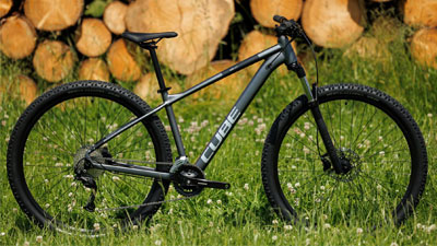 Se ve una bicicleta de montaña Cube gris, parada en un prado. Al fondo hay madera apilada.