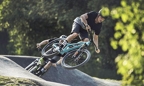 Ciclista saltando con una bicicleta de cross azul claro en un bike park, otro ciclista con una bicicleta verde al fondo.