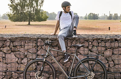 Se ve a un hombre con casco sentado en un muro. En primer plano hay una bicicleta gris y al fondo se ve un paisaje campestre.
