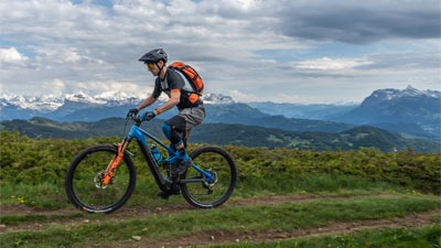 Se ve a un hombre montado en una bicicleta eléctrica Cube Stereo Hybrid azul con detalles naranjas. Va por un sendero y al fondo se ven montañas.