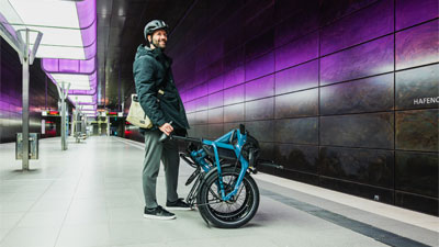 Se ve a un hombre en una estación de metro, delante de él hay una bicicleta plegable azul.