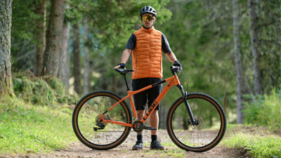 Un hombre está detrás de su bicicleta de montaña naranja Cube, en un camino forestal.