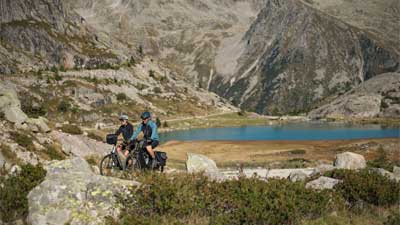 Una pareja recorre en bicicleta de montaña un paisaje pedregoso, con montañas y un lago al fondo.