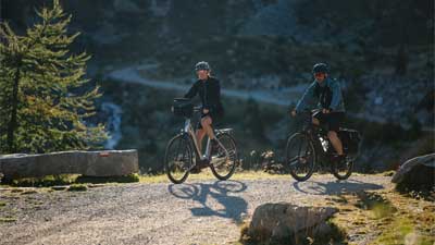 Una pareja va en bicicleta de montaña por un carril bici, con un paisaje montañoso al fondo.