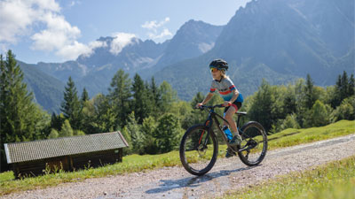 Un niño va en su bicicleta de montaña por un camino de grava a través de un paisaje montañoso. Al fondo se ve una cabaña.