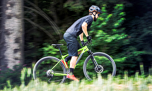 Ciclista de pie sobre una bicicleta de montaña rígida verde, circulando por un entorno natural verde.