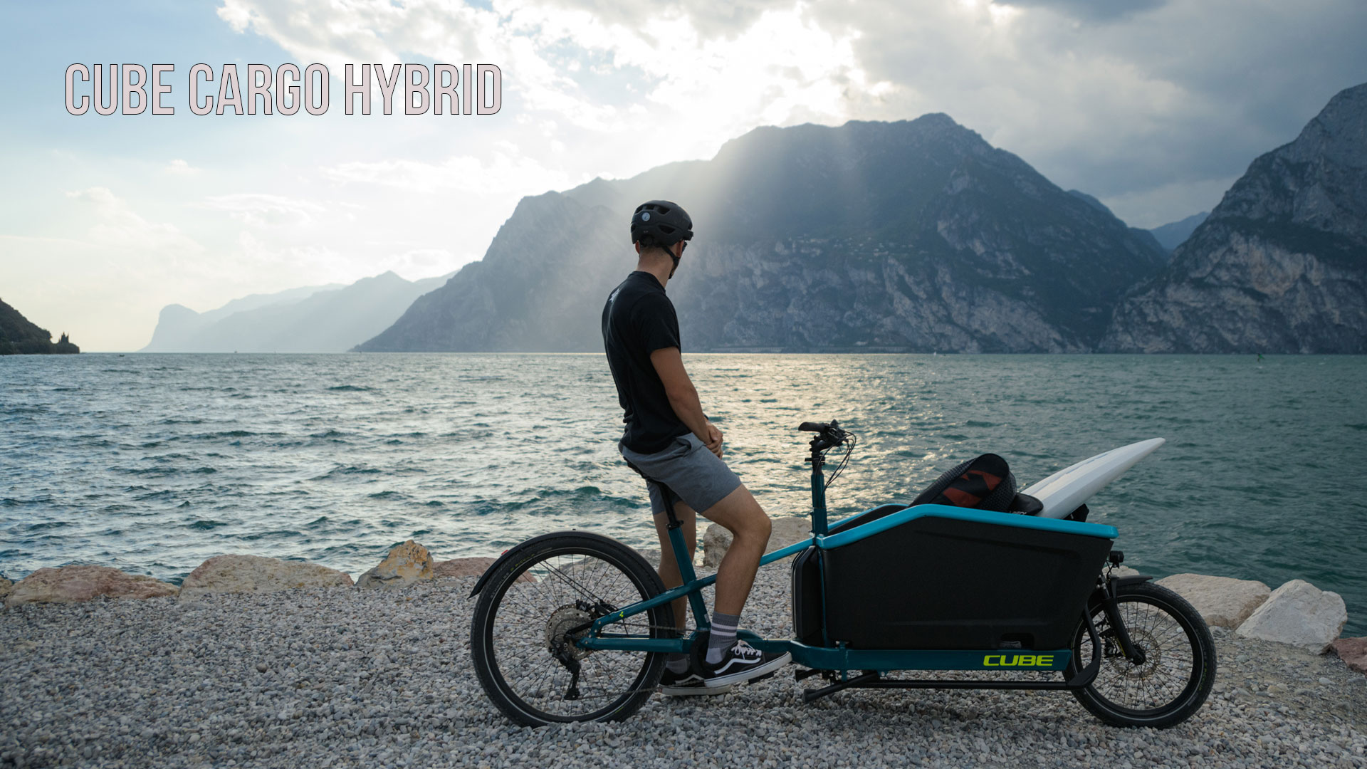 Se ve a un hombre en una bicicleta de carga azul, parado a la orilla de un lago y contemplando las montañas al fondo. La imagen lleva por título «Cube Cargo Hybrid».