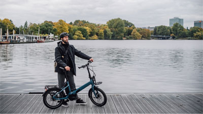 Un hombre está de pie junto a su bicicleta plegable azul en un embarcadero. Al fondo se ve el agua y una ciudad.