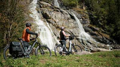 Una pareja empuja sus bicicletas por un prado, con una cascada al fondo.