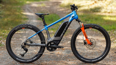 Se ve una bicicleta eléctrica para niños Cube de color gris con detalles en azul y naranja. Está parada en un camino forestal.