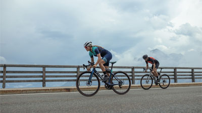 Se ven dos ciclistas de carretera, con una valla de madera y nubes al fondo.