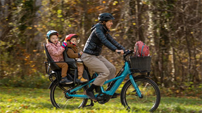 Se ve a una madre en una bicicleta eléctrica de carga Cube Longtail Hybrid azul. En el portaequipajes van sentados dos niños pequeños.